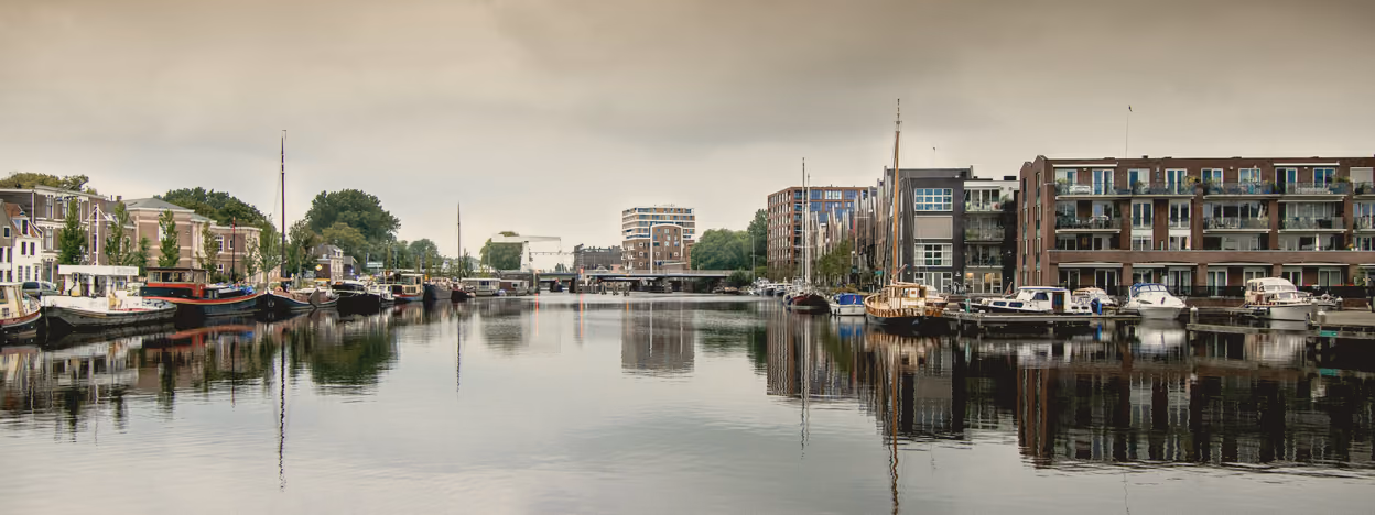 The Spaarne at Haarlem under a flat pale sky, barges moored along both banks and a bridge in the distance.