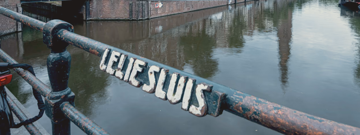A weathered iron bridge railing on the Prinsengracht with the sluice name painted along it in white capitals, the canal running away behind.