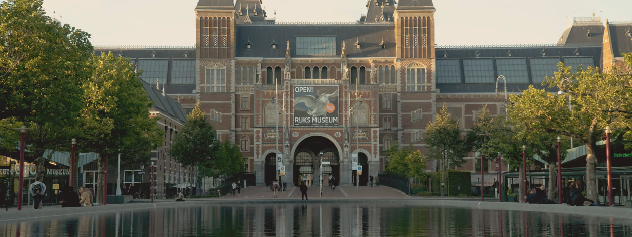 The Rijksmuseum seen across the shallow pond on the Museumplein, its facade reflected in the water under a flat sky.