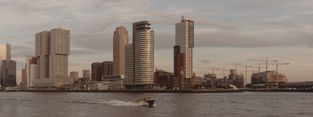 The Rotterdam skyline across the Nieuwe Maas seen low from the water, with construction cranes at the right and a small boat crossing.